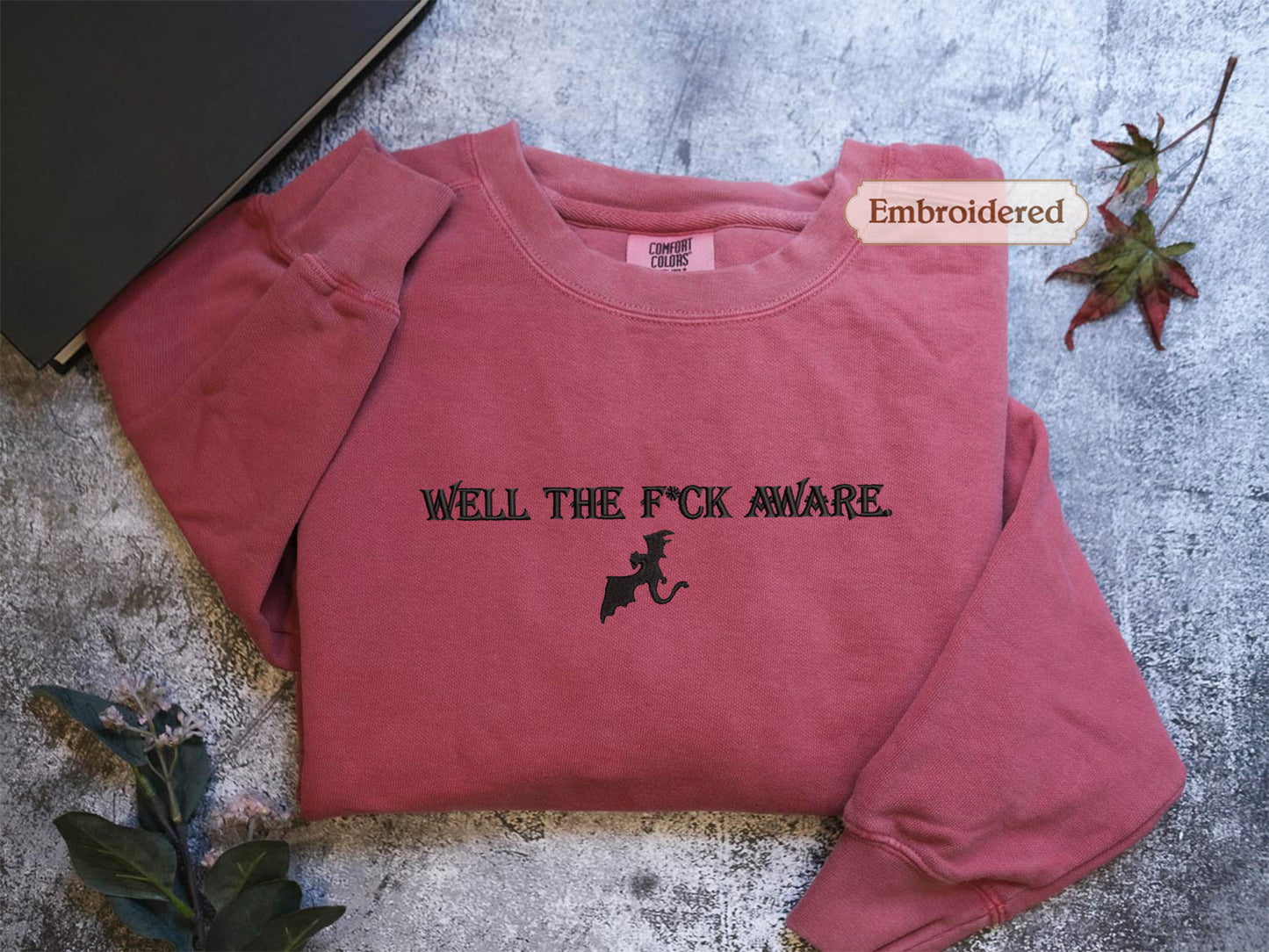 A red sweatshirt with the text "WELL THE F*CK AWARE" printed on it, along with a graphic of a cat. The sweatshirt is placed on a gray surface, and there is a leaf and a book visible in the background.
