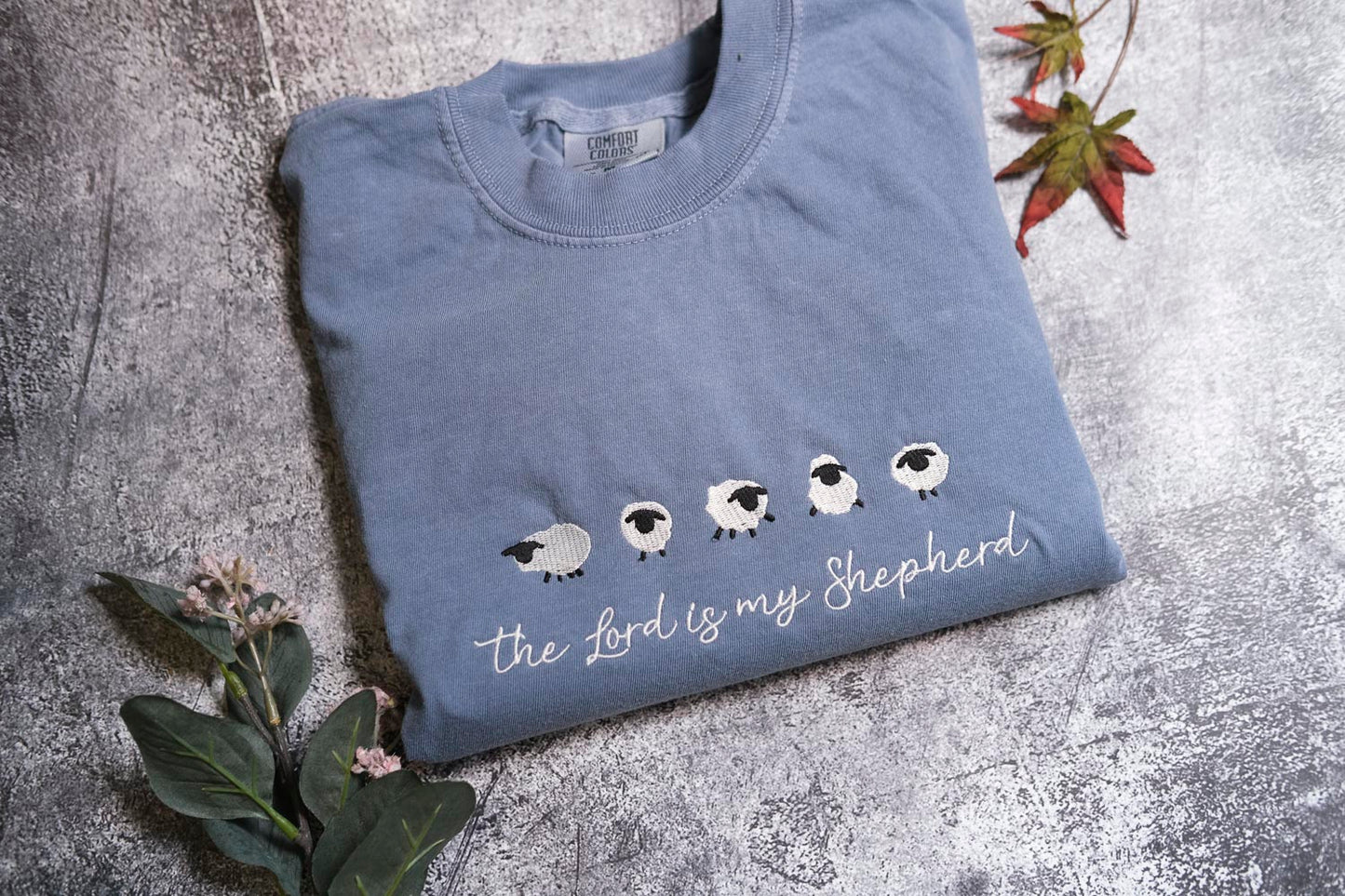 A blue t-shirt with a white sheep embroidered design and the text "The Lord is my Shepherd" printed on it, along with a small leaf and a flower.
