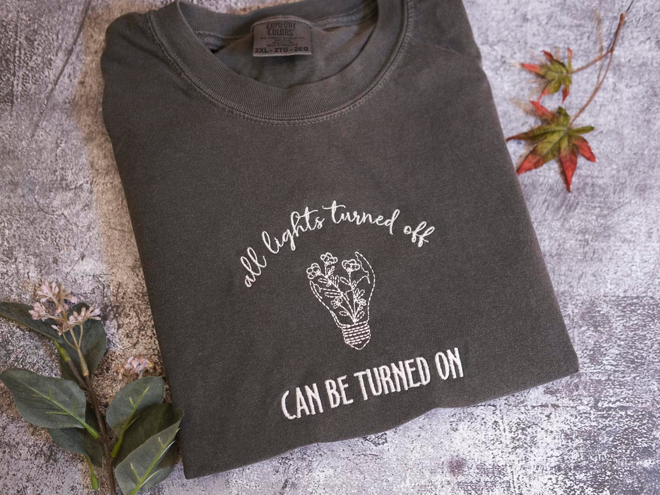 A gray t-shirt with the text "all lights turned off" and a light bulb graphic, along with the phrase "can be turned on" printed on it, is displayed on a gray surface.