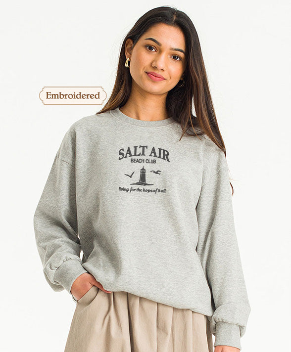 A young woman wearing a gray sweatshirt with the text "Salt Air Beach Club" printed on it, standing against a white background.
