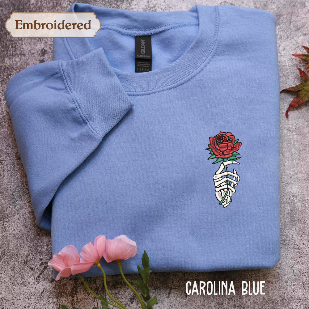 a blue long-sleeved shirt with a rose embroidered on the front, along with a pair of pink flowers. The shirt is laid out on a gray surface, and there is a label visible on the collar.