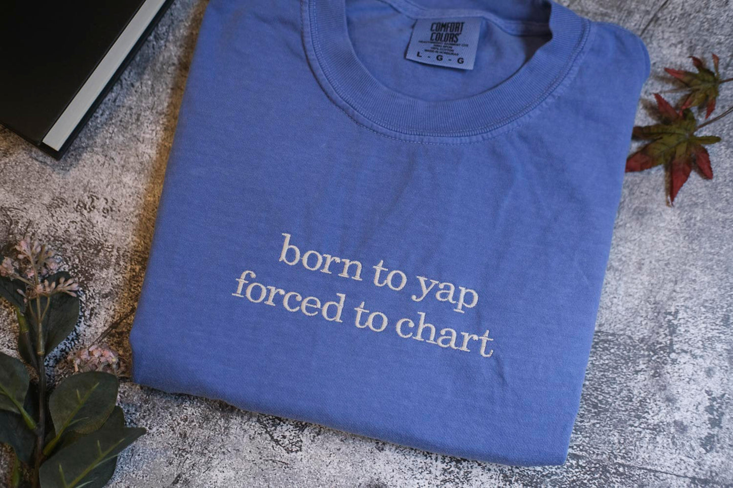 A blue t-shirt with the text "born to yap forced to chart" printed on it, along with a label indicating it is from the "COMPACT COLOR'S" brand. The t-shirt is placed on a surface with some leaves and a book, creating a casual, laid-back atmosphere.