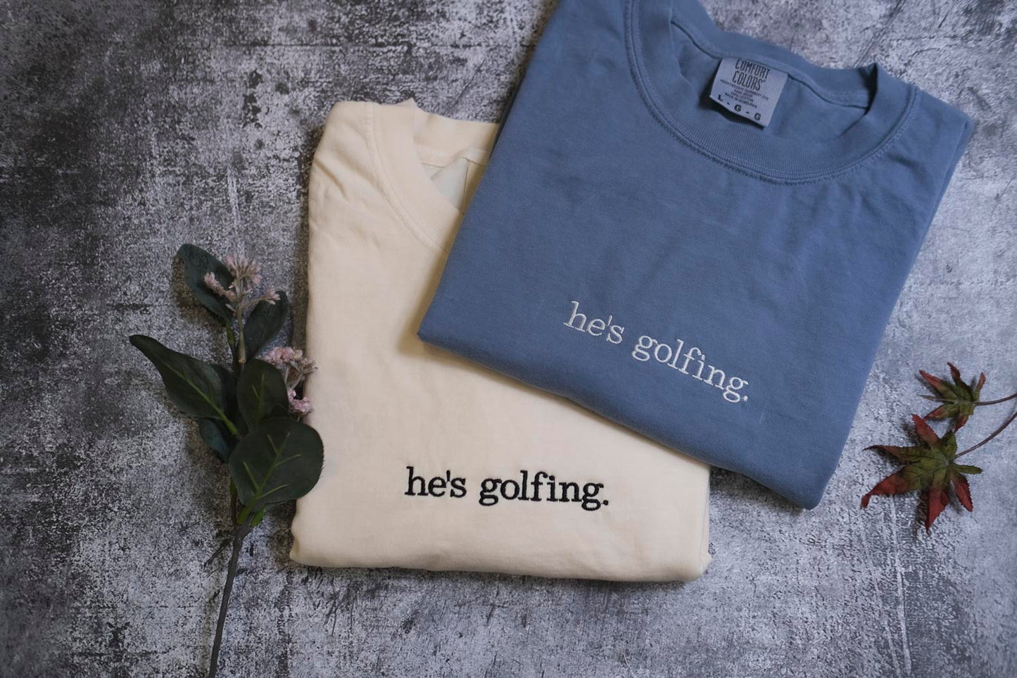 two t-shirts with the text "he's golfing" printed on them, along with some greenery and a leaf.