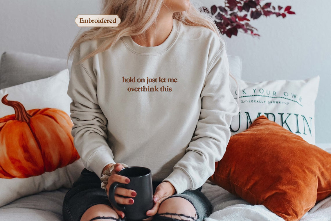 A woman sitting on a bed with a white sweater that says "hold on just let me overwhelm this" embroidered on it. She is holding a black mug and surrounded by pillows and a pumpkin-themed pillow.