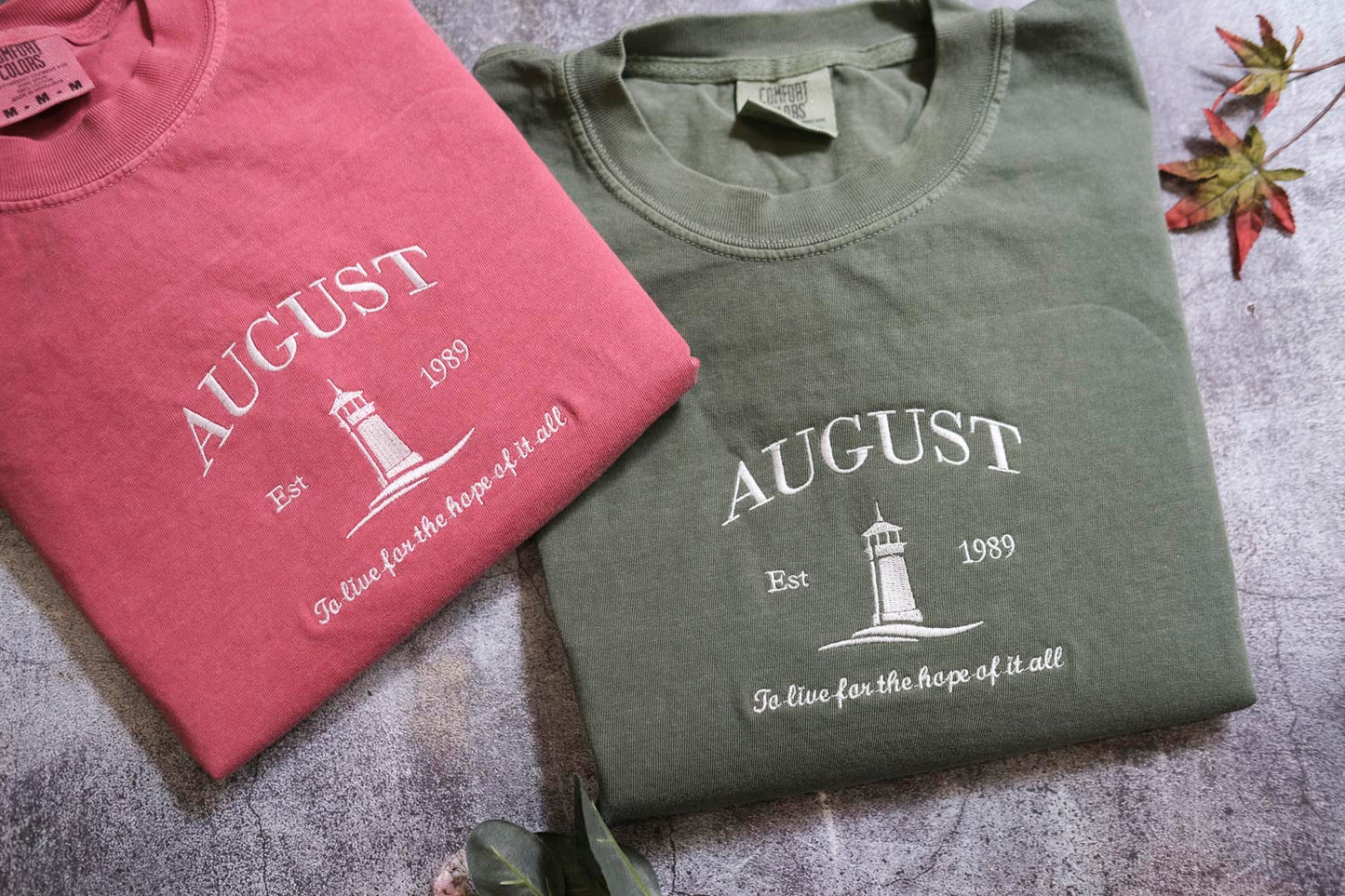 two t-shirts with the word "AUGUST" printed on them, along with a lighthouse graphic. The t-shirts are laid out on a surface, and there are some leaves and a plant visible in the background.
