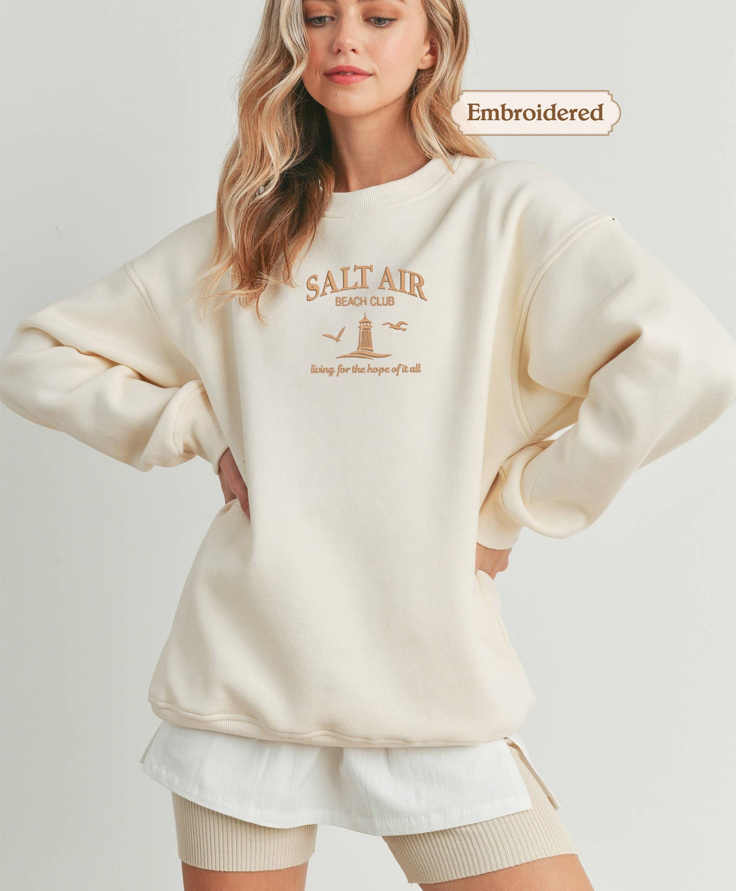 A woman wearing a cream-colored sweater with the text "Salt Air Beach Club" printed on it, standing with her hands on her hips.