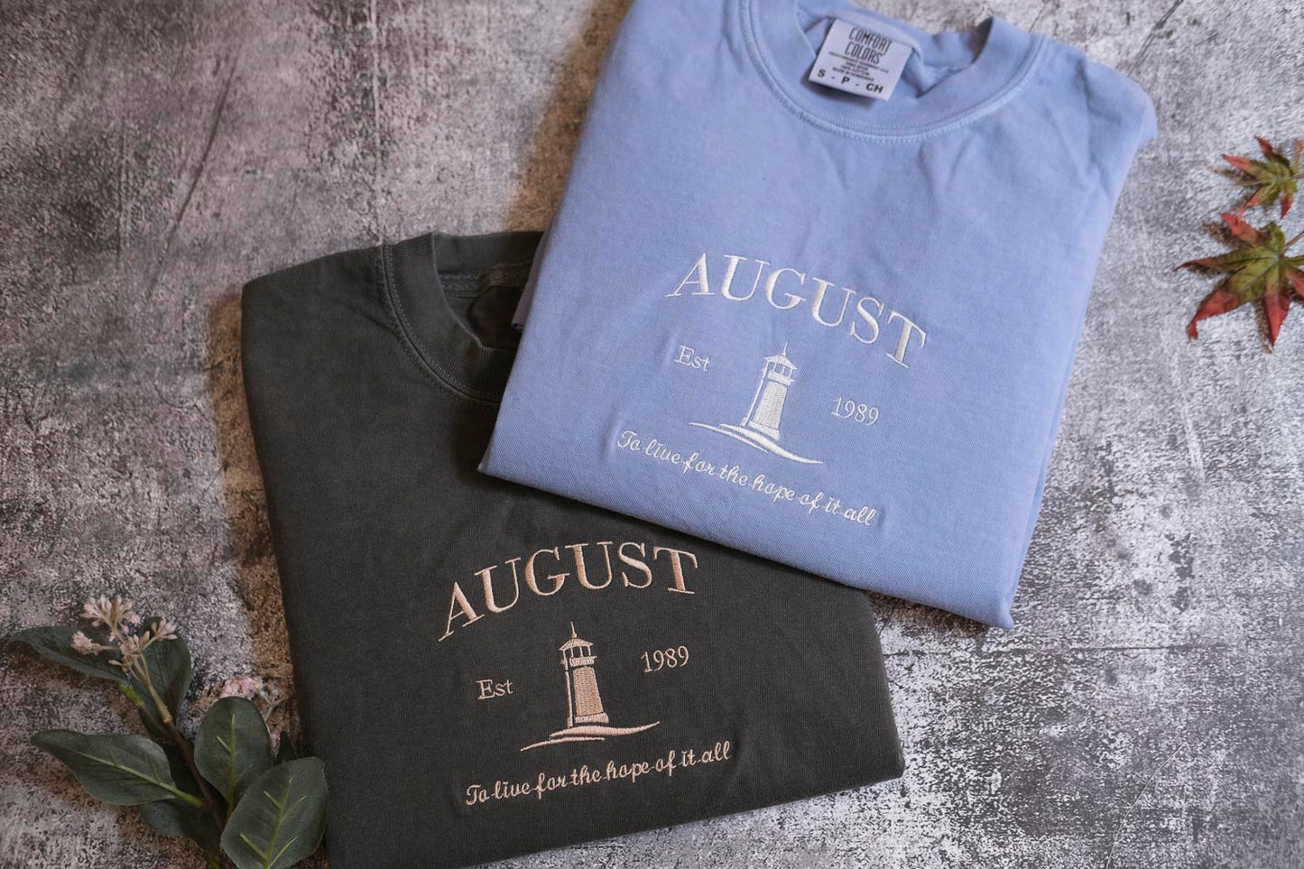 two t-shirts with the word "AUGUST" printed on them, laid out on a textured surface.