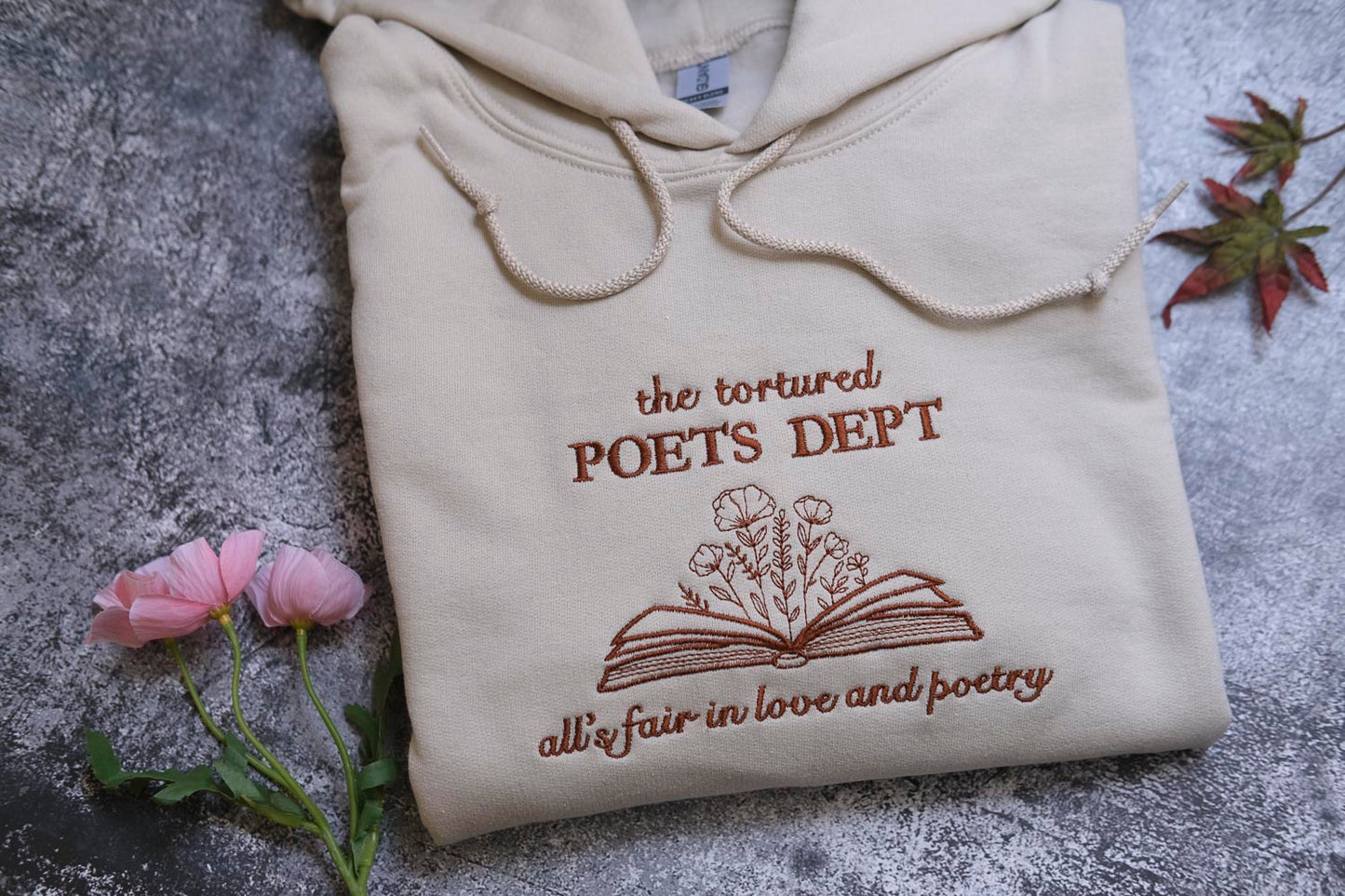 a beige hoodie with a brown embroidered design, which says "the tortured POET'S DEPT all fair in love and poetry". The hoodie is placed on a gray surface, with a small bouquet of pink flowers and a fallen leaf nearby.