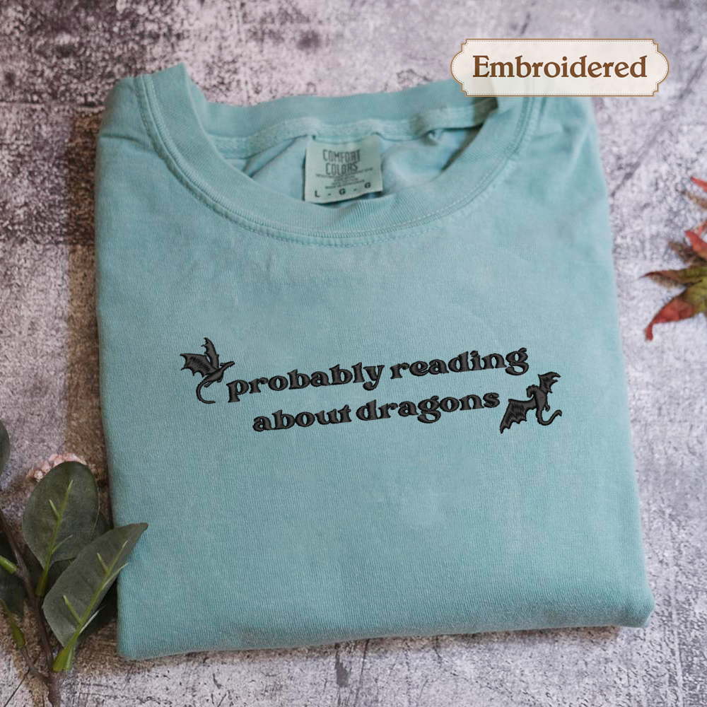 A light blue t-shirt with the text "probably reading about dragons" embroidered on it, along with a small dragon design.