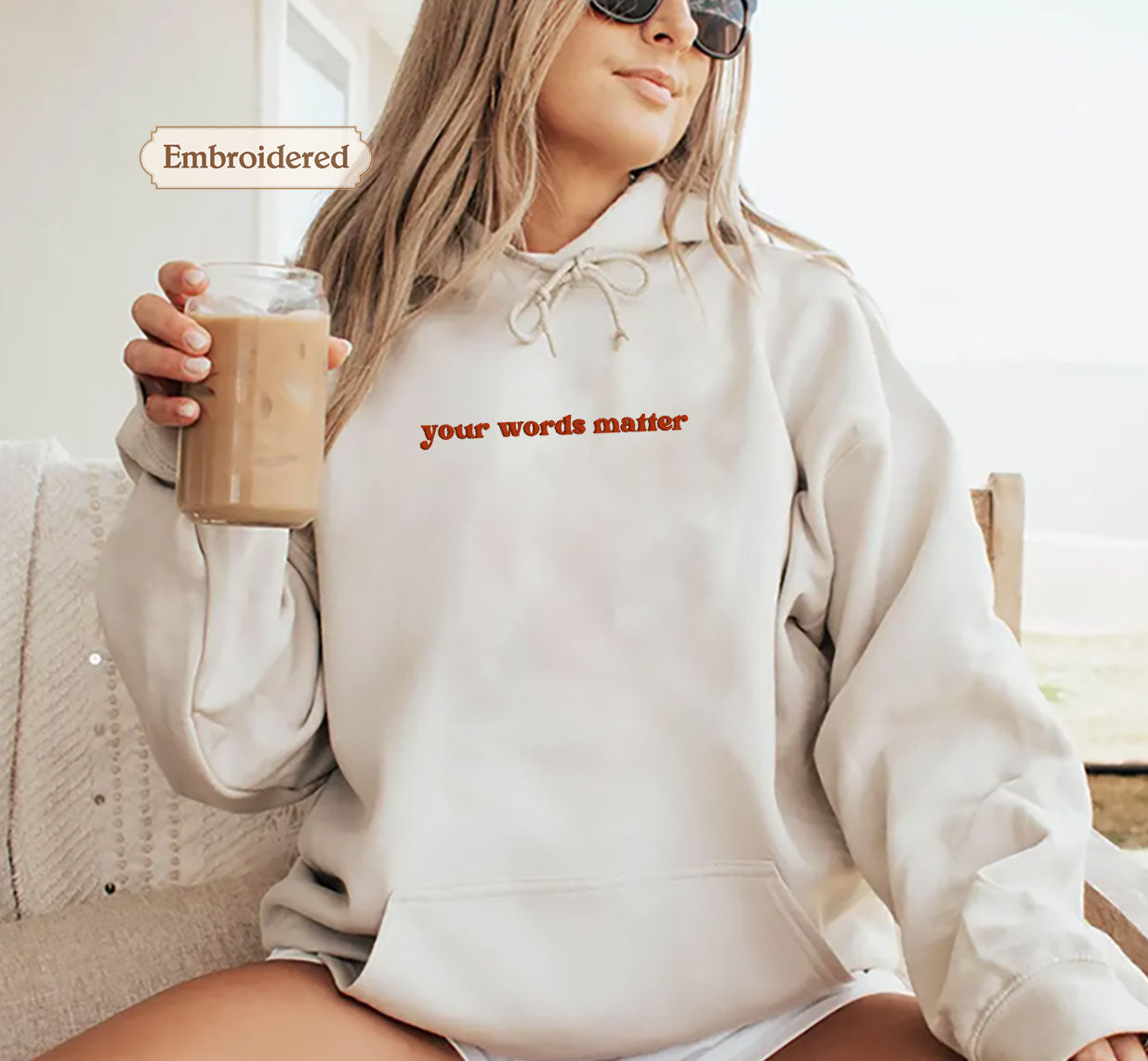 A woman wearing a white hoodie with the text "your words matter" is sitting on a couch, holding a glass of what appears to be iced coffee.