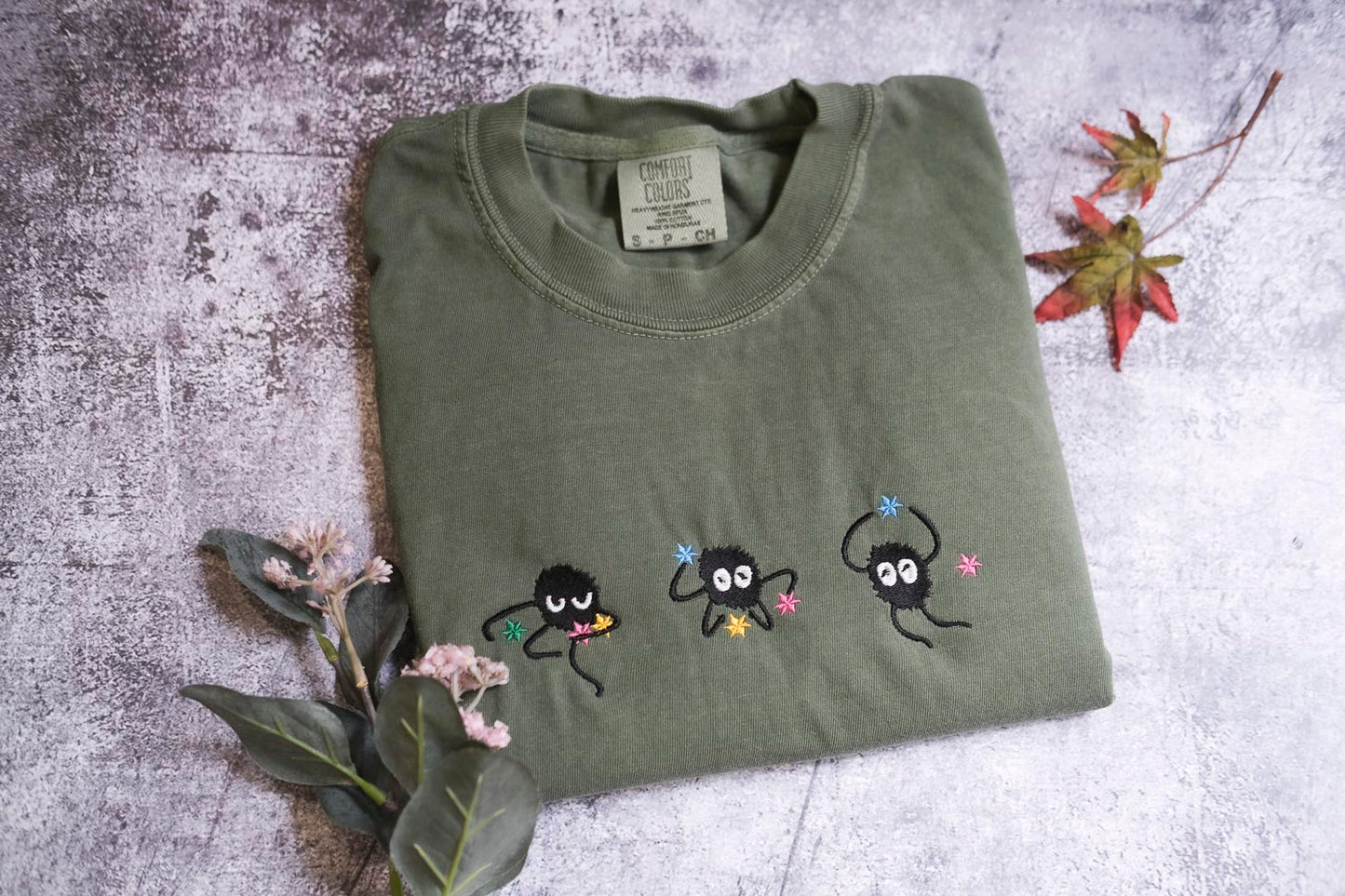 a green t-shirt with three small black figures and flowers embroidered on it, along with some leaves and flowers on a textured surface.