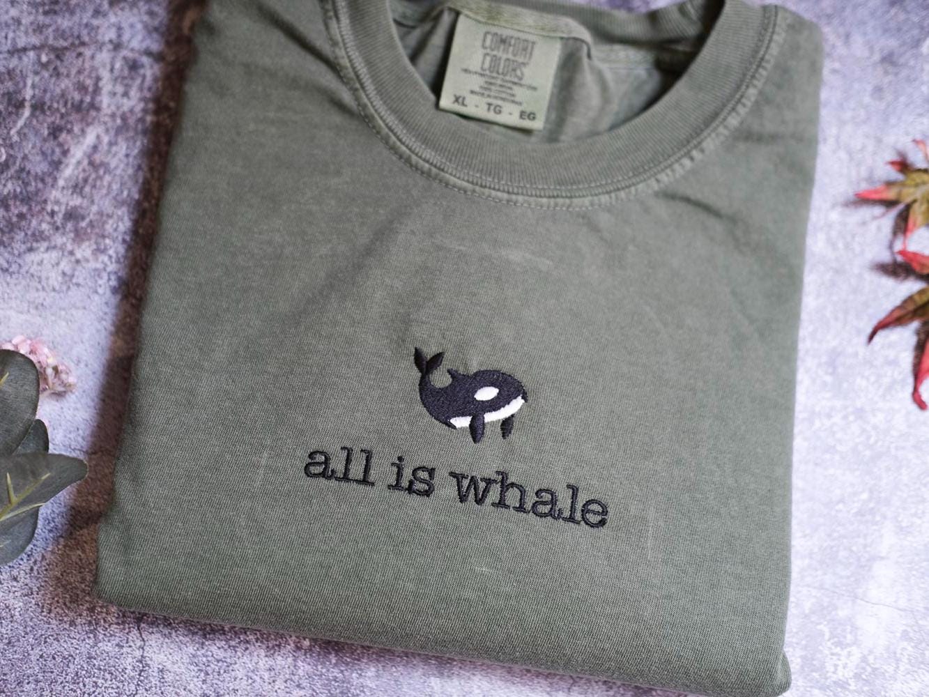 a gray t-shirt with a black and white whale embroidered on it, along with the text "all is whale" printed on the front.