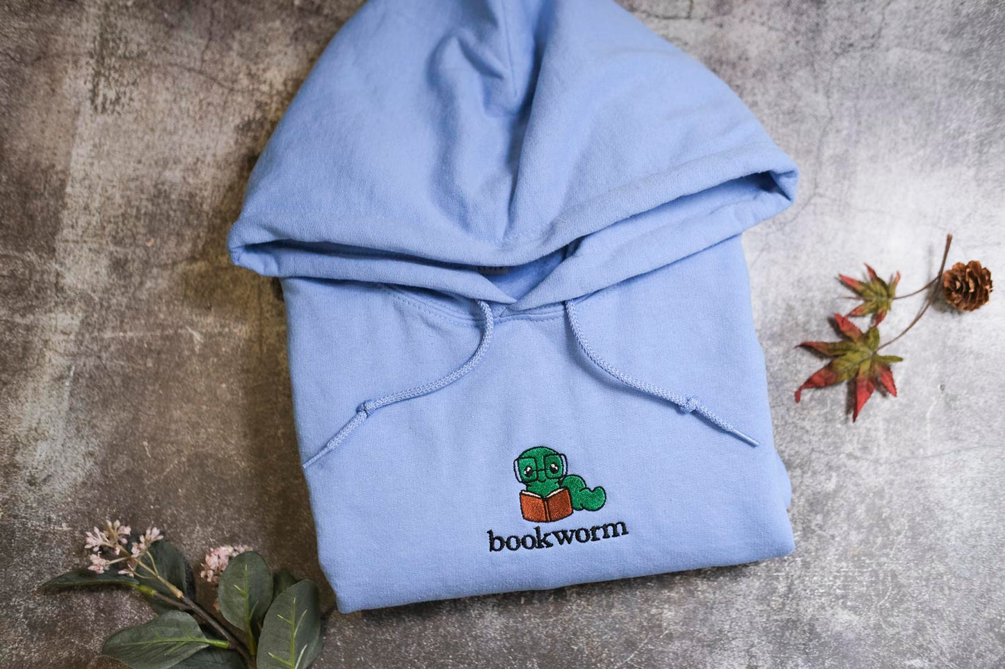 A light blue hoodie with a cartoon image of a caterpillar reading a book, along with the text "bookworm" printed on it, is laid out on a gray surface. There are some leaves and a pine cone nearby, and a small flower is also visible.