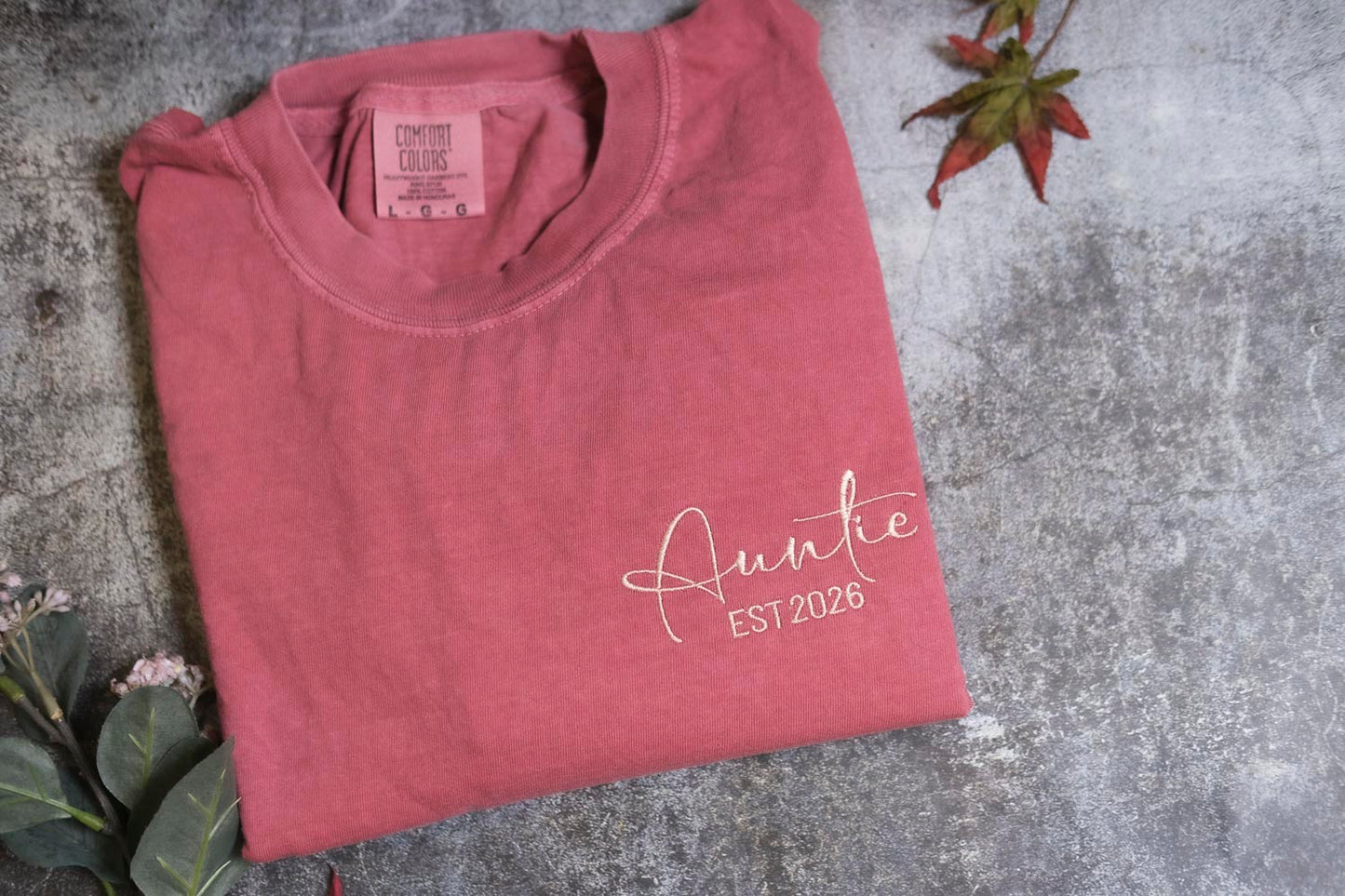 A pink t-shirt with the text "Auntie EST2026" printed on it, along with a small label on the collar. The t-shirt is placed on a gray surface, with a small plant and a red leaf visible in the background.