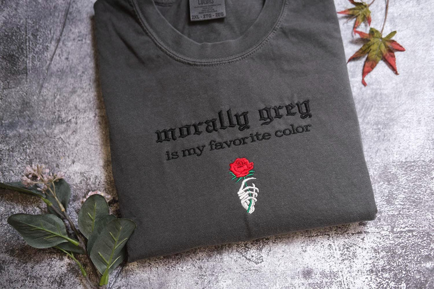 a gray t-shirt with a red rose and a skeleton hand embroidered on it, along with some leaves and a stem.
