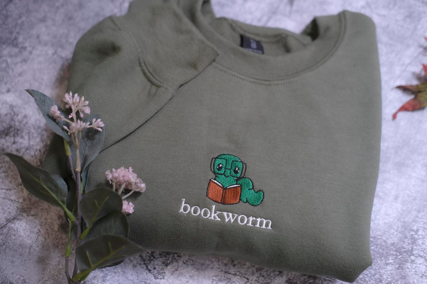A green sweatshirt with a cartoon image of a bookworm and the word "bookworm" printed on it, along with some pink flowers and a leaf.