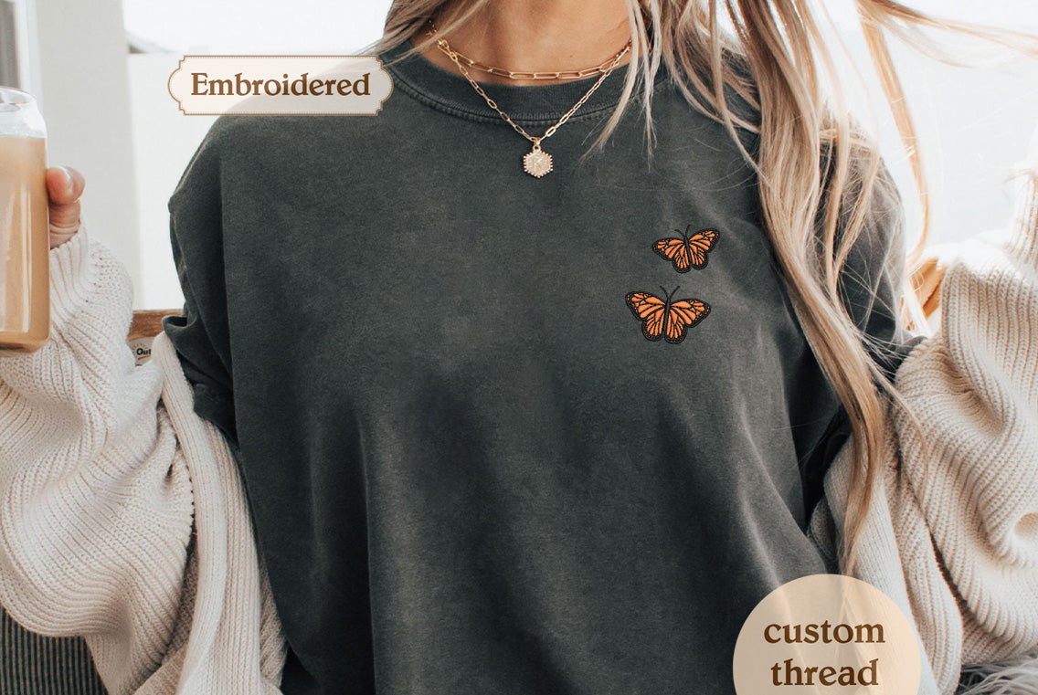 A person wearing a gray t-shirt with two embroidered butterflies on the front, along with a necklace and a sweater.
