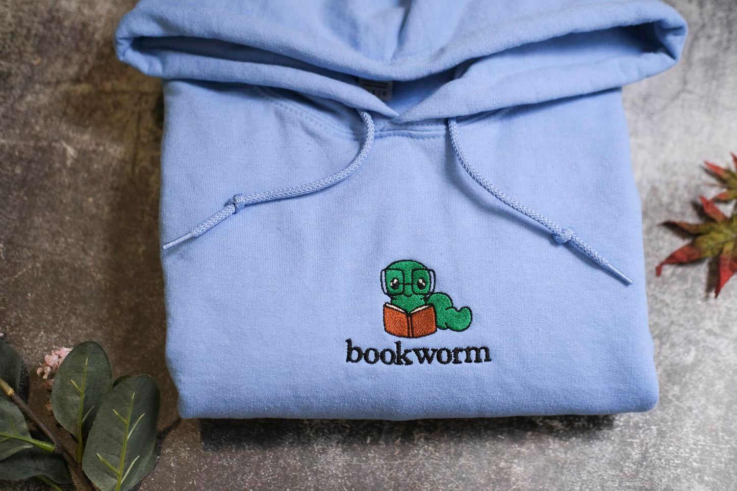 A light blue hoodie with a cartoon image of a book worm and the text "bookworm" printed on it, along with some leaves and a small plant.