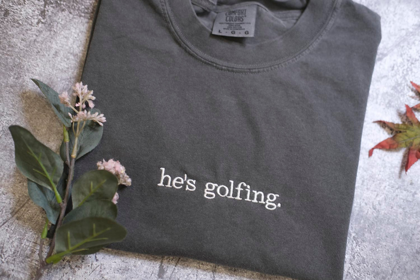 A gray t-shirt with the text "he's golfing" printed on it, along with some flowers and a leaf on the side.