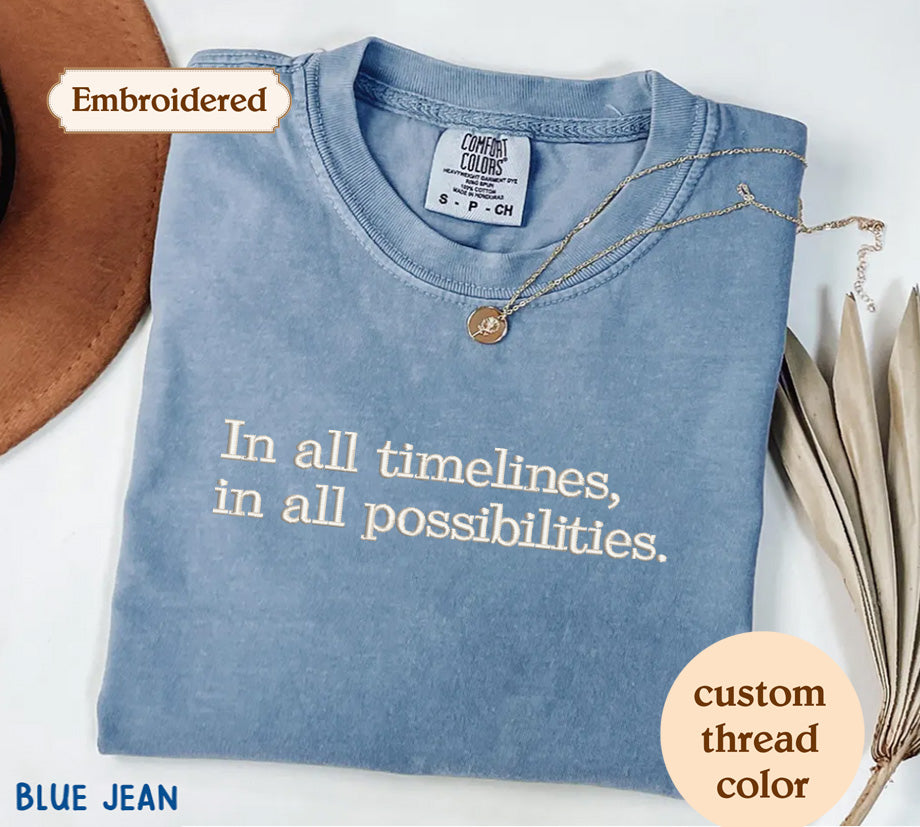 a blue t-shirt with the text "In all timelines, in all possibilities." The t-shirt is embroidered with the text and is accompanied by a necklace and some leaves.