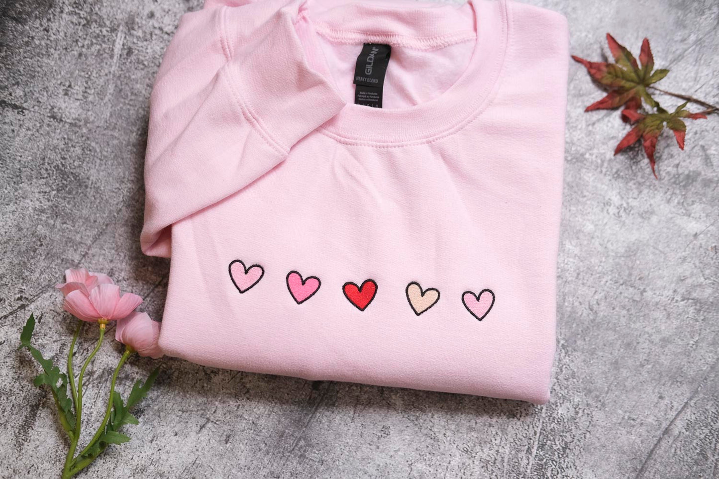 A pink sweatshirt with a row of five hearts embroidered on it, along with a single pink rose and a small red leaf.