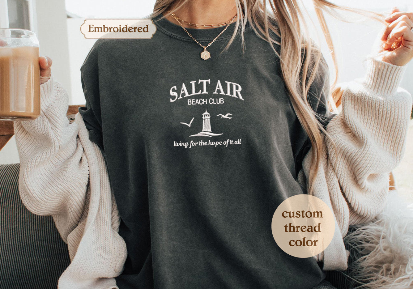 A person wearing a gray t-shirt with the text "Salt Air Beach Club" printed on it, along with a lighthouse and birds. The person is also wearing a white sweater and holding a drink.