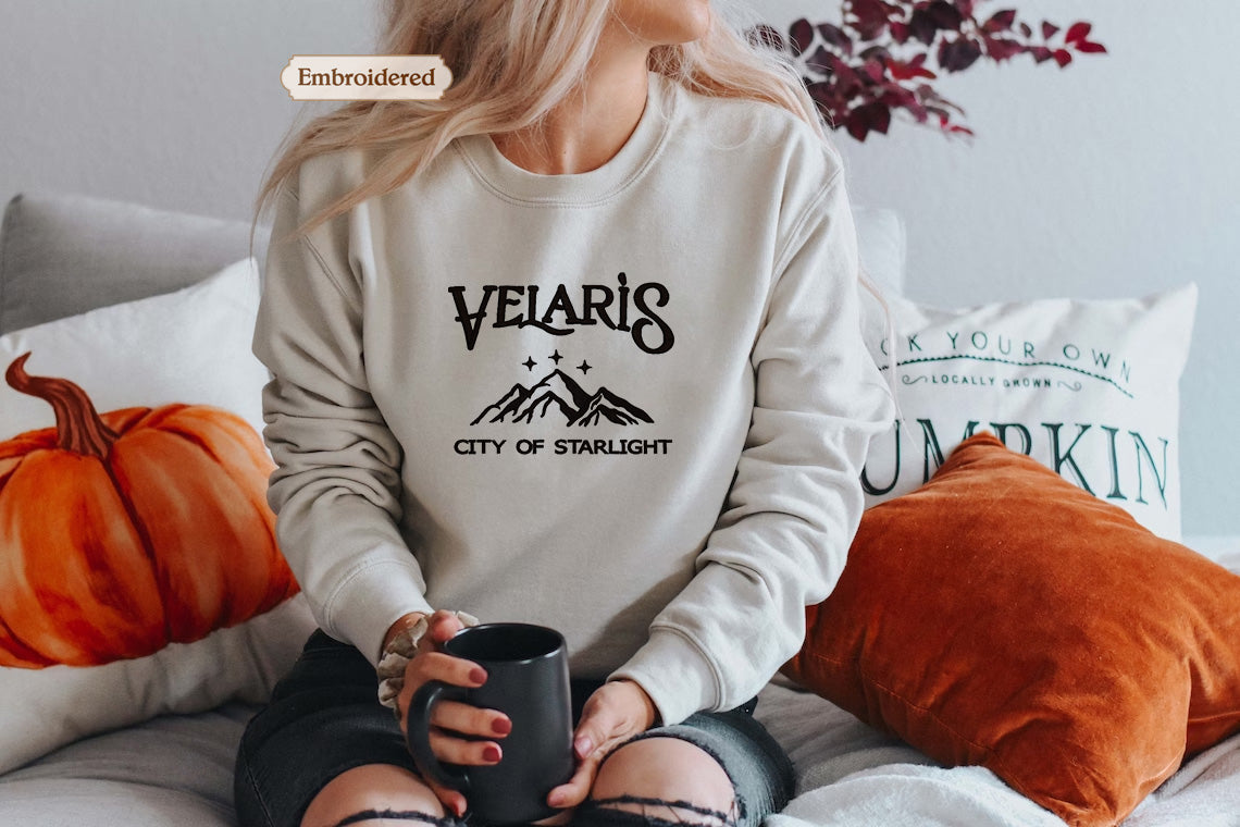 A woman wearing a white sweatshirt with the text "VELARIIS CITY OF STARLIGHT" sits on a bed with pillows and a pumpkin pillow. She is holding a black mug in her hand.