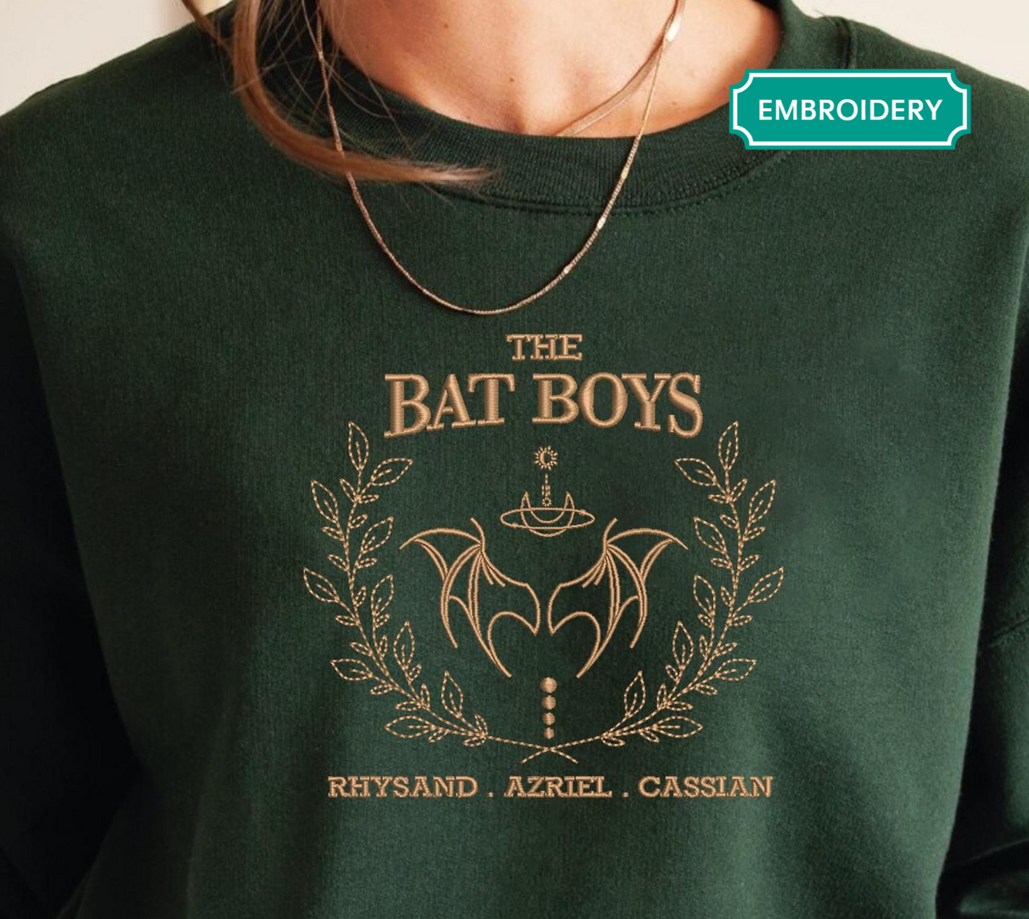 A green sweater with a gold embroidered design featuring the text "THE BAT BOYS" and the names " RHYSAND, AZRIEL, CASSIAN" around a bat emblem.