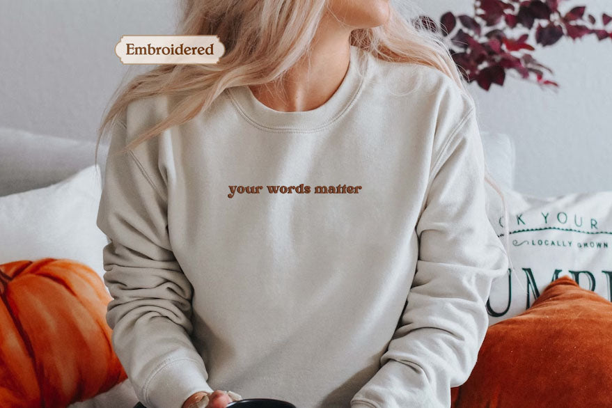 A woman wearing a white sweatshirt with the text "your words matter" embroidered on it, sitting on a couch with orange pillows.
