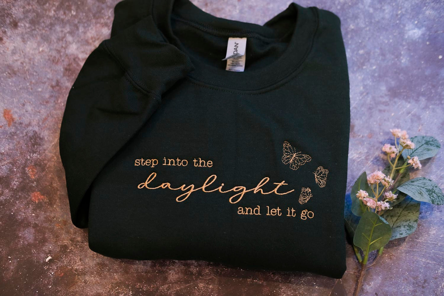 A black sweatshirt with gold text and butterflies printed on it, placed on a textured surface alongside a small plant with white flowers.