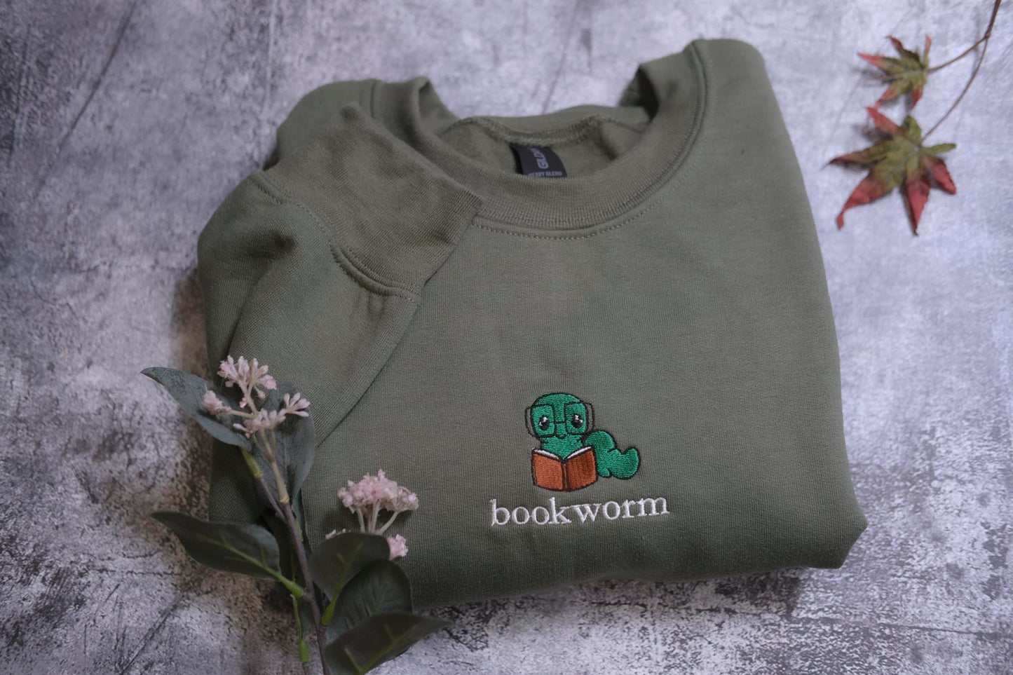 A green sweatshirt with a cartoon image of a caterpillar reading a book, along with the text "bookworm" printed on it, is displayed on a gray surface alongside some pink flowers.