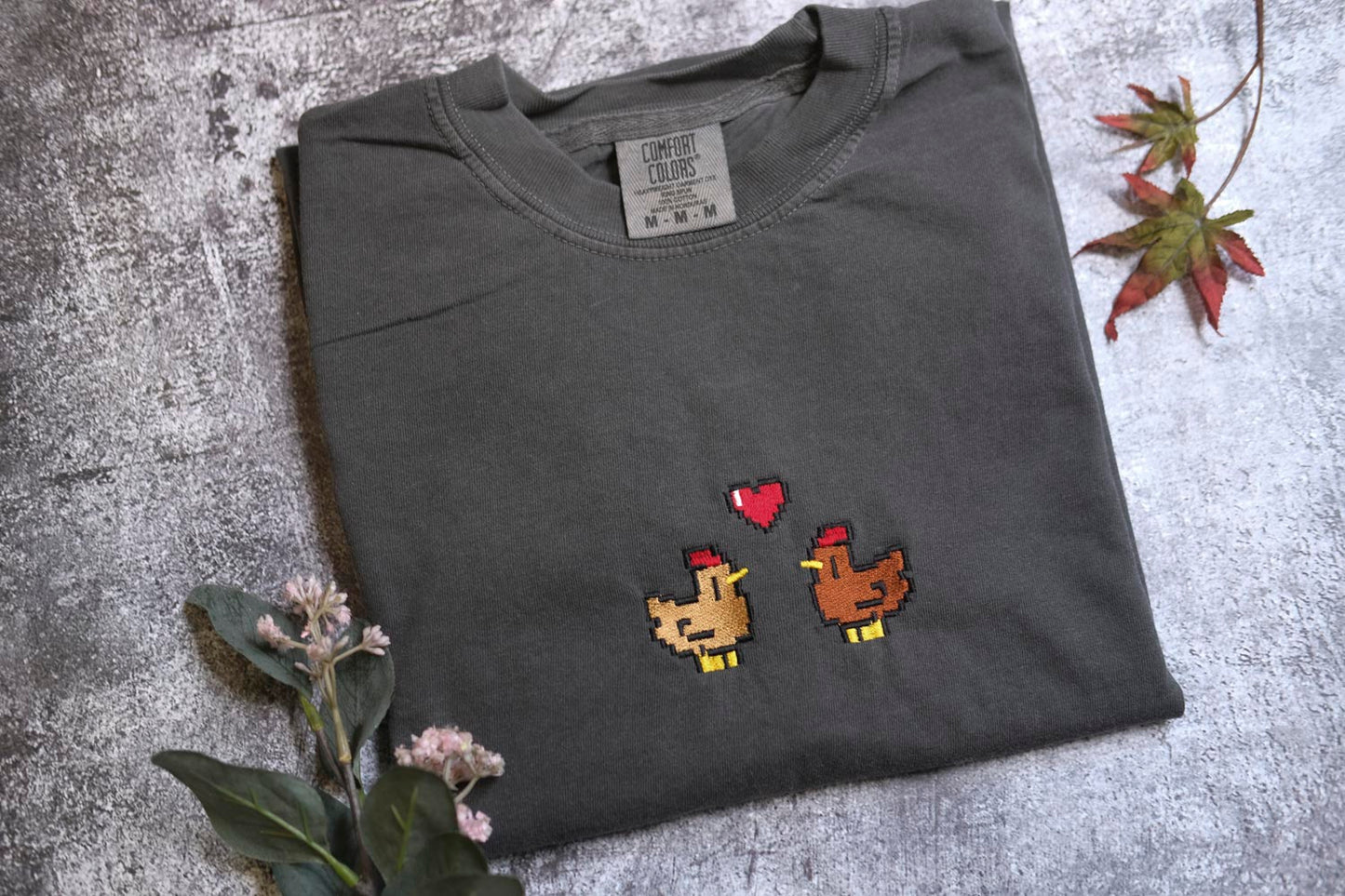 a gray t-shirt with a pixelated design of two chickens, one brown and one yellow, with a heart symbol between them. The t-shirt is placed on a gray surface, and there are some flowers and a leaf visible in the background.