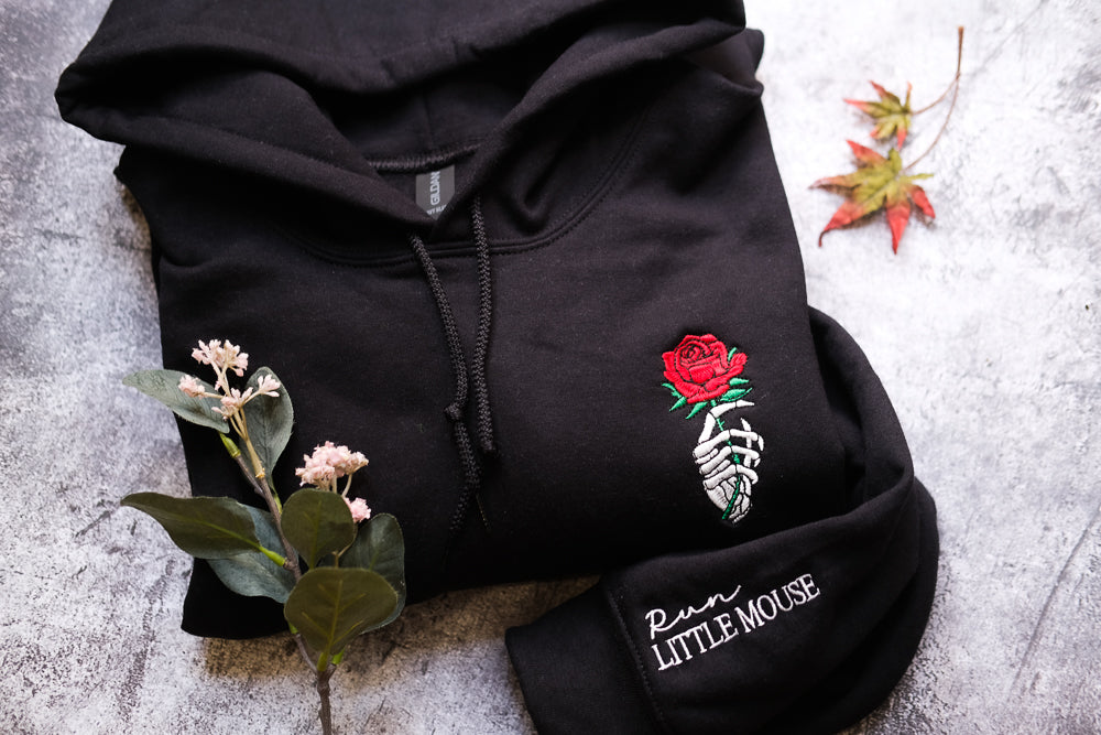 a black hoodie with a red rose embroidered on the front, along with a small plant and a red leaf.