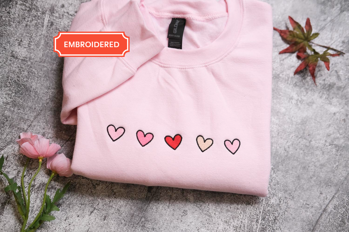 A pink t-shirt with a row of five hearts embroidered on it, along with a small red heart and a pink rose, placed on a gray surface.