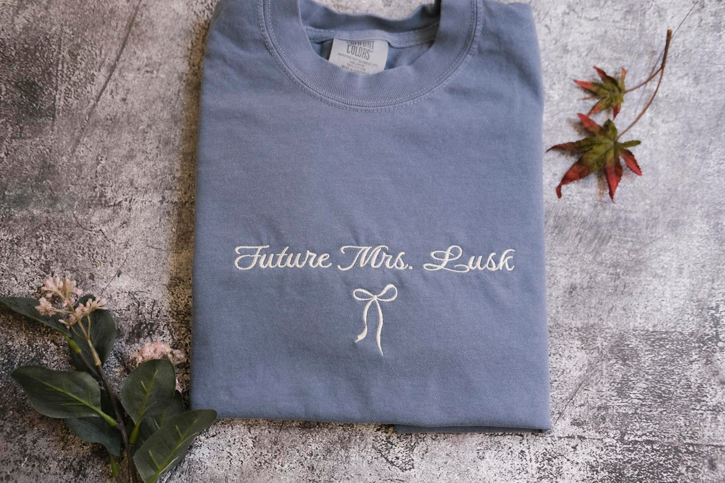 A gray t-shirt with the text "Future Mrs. Lush" printed on it, along with a bow symbol, is displayed on a gray surface alongside some leaves and flowers.