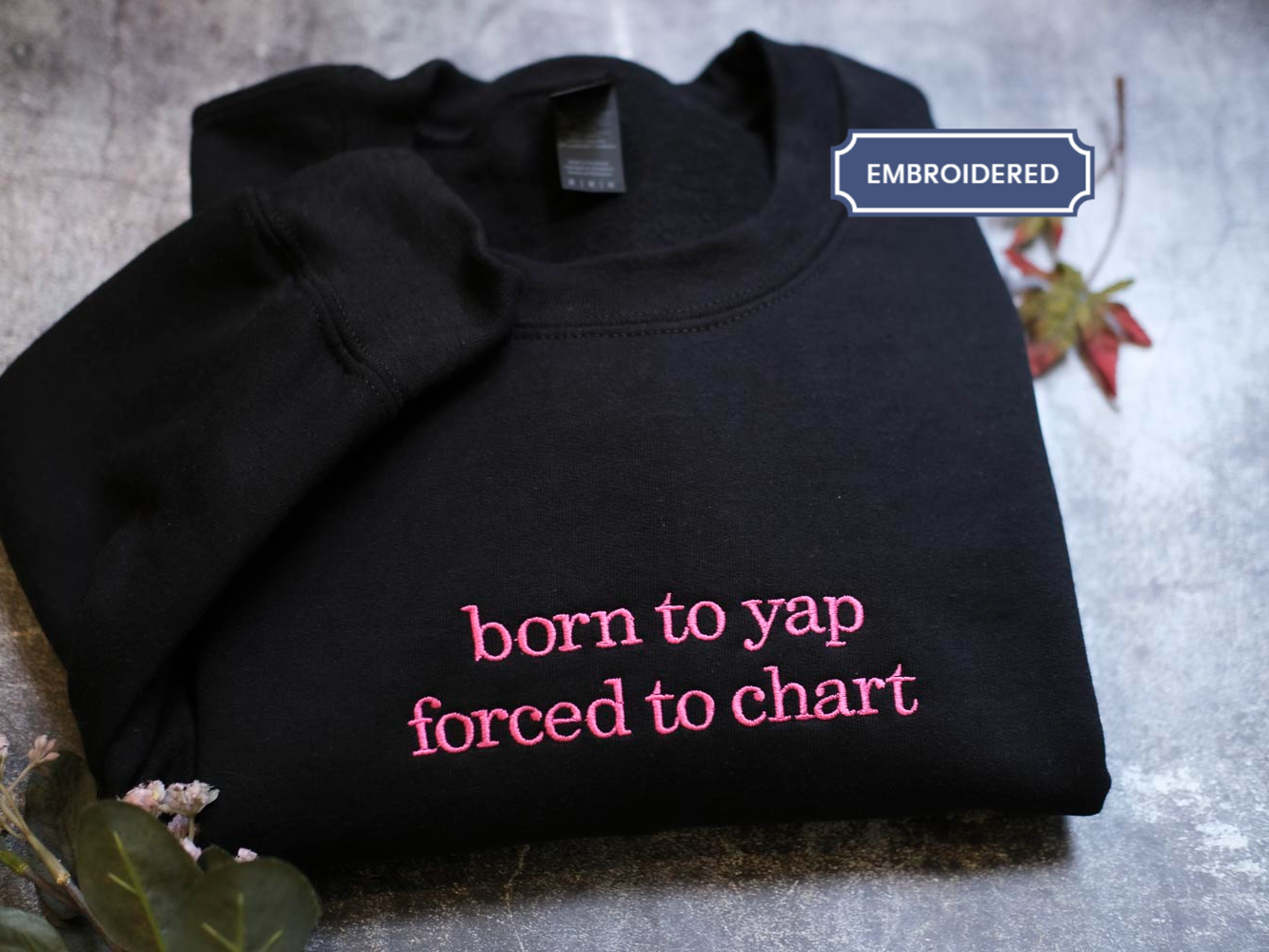A black sweatshirt with the text "born to yap forced to chart" printed on it, along with a small pink flower and a leaf.
