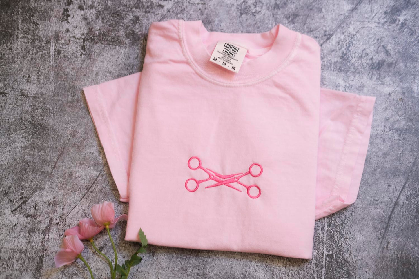 A pink t-shirt with a pink scissors design is displayed on a gray surface, accompanied by a small bouquet of pink flowers.