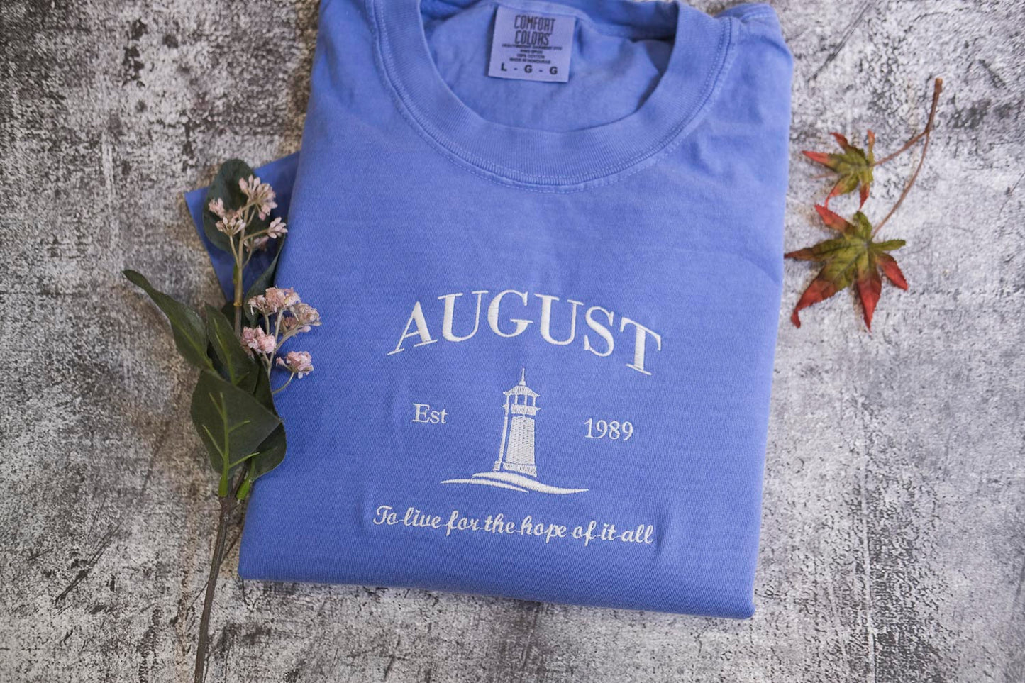 A blue t-shirt with the text "AUGUST Est 1989 To live for the hope of it all" printed on it, along with a lighthouse graphic, is displayed on a textured surface alongside some flowers and a fallen leaf.