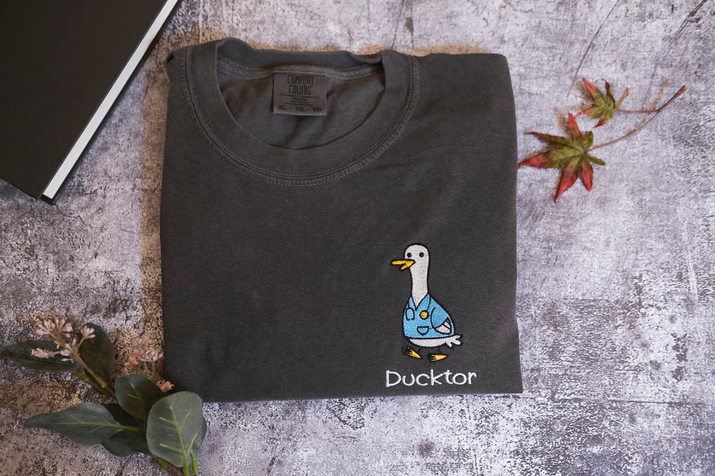 a gray t-shirt with a cartoon duck wearing a blue shirt and the word "Duckster" printed on it, along with a small leaf and a book.