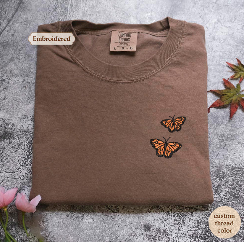 a brown t-shirt with a label and two orange butterflies embroidered on the front. The t-shirt is placed on a gray surface, and there are some leaves and flowers in the background.