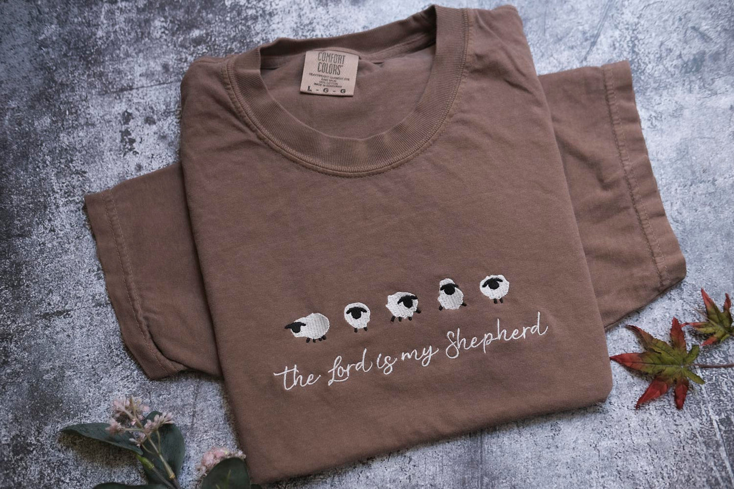 A brown t-shirt with a printed design of sheep and the text "The Lord is my Shepherd" is displayed on a gray surface, accompanied by a small leaf and some flowers.