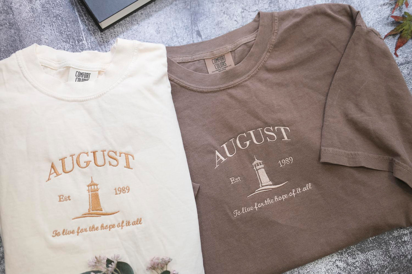 two t-shirts with the word "AUGUST" printed on them, along with a lighthouse and the text "Est. 1989" and "To live for the hope of it all." The t-shirts are laid out on a surface, with a book visible in the background.