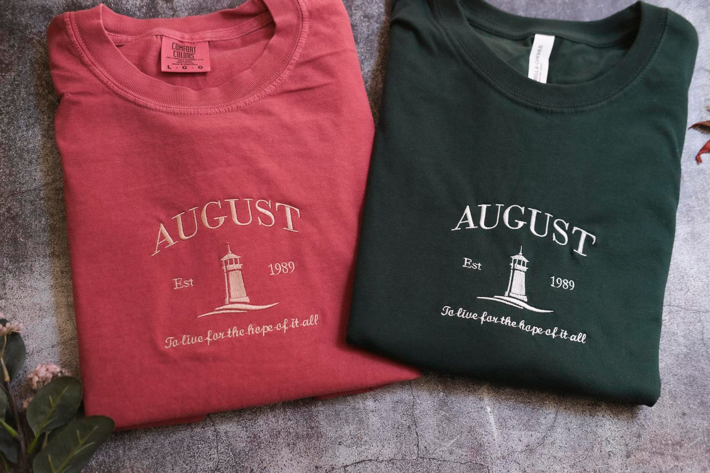 two t-shirts with the word "AUGUST" printed on them, along with the text "Est. 1989" and "To live for the hope of it all" printed below. The t-shirts are laid out on a gray surface, with some leaves visible in the background.