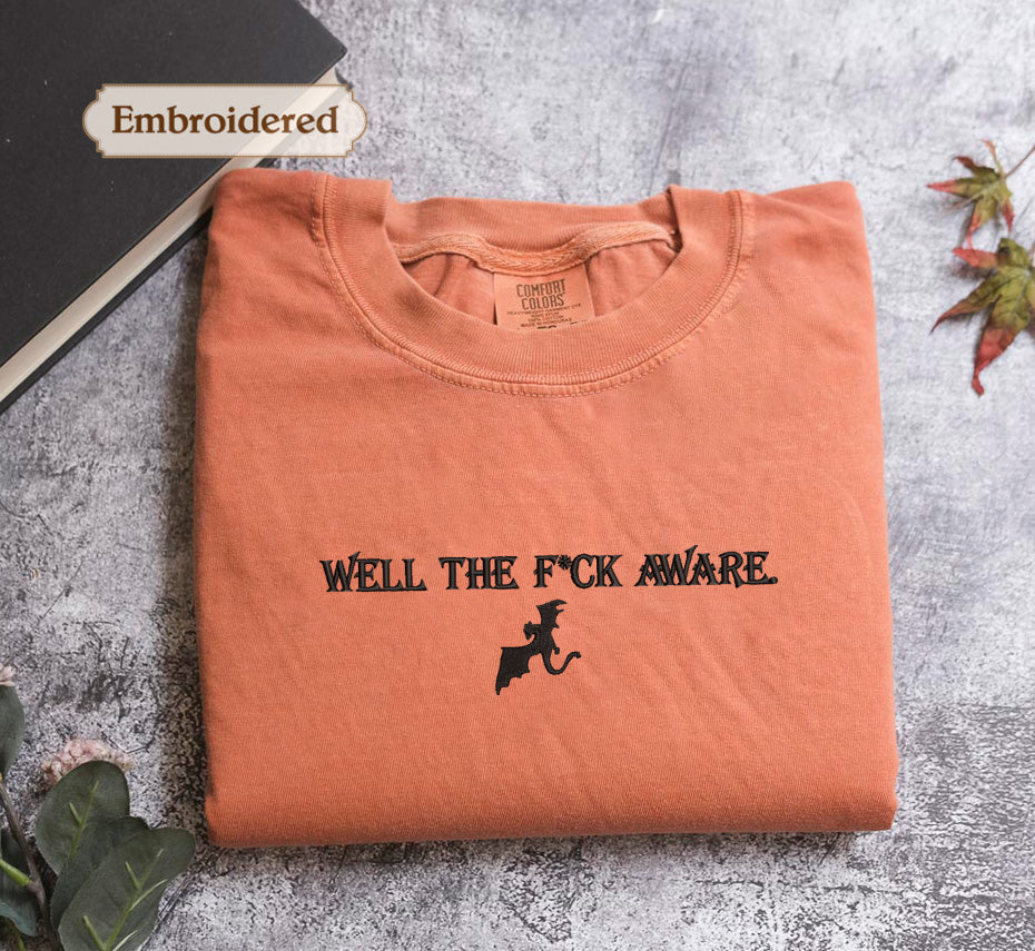 a folded orange t-shirt with the text "WELL THE F*CK AWARE" printed on it, along with a graphic of a bat. The t-shirt is placed on a gray surface, and there is a book and some leaves in the background.