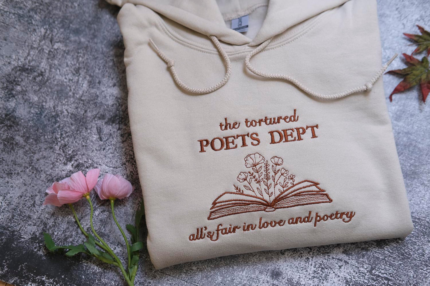 a beige hoodie with a brown embroidered design, including text and an open book with flowers, placed on a gray surface alongside a few pink flowers.