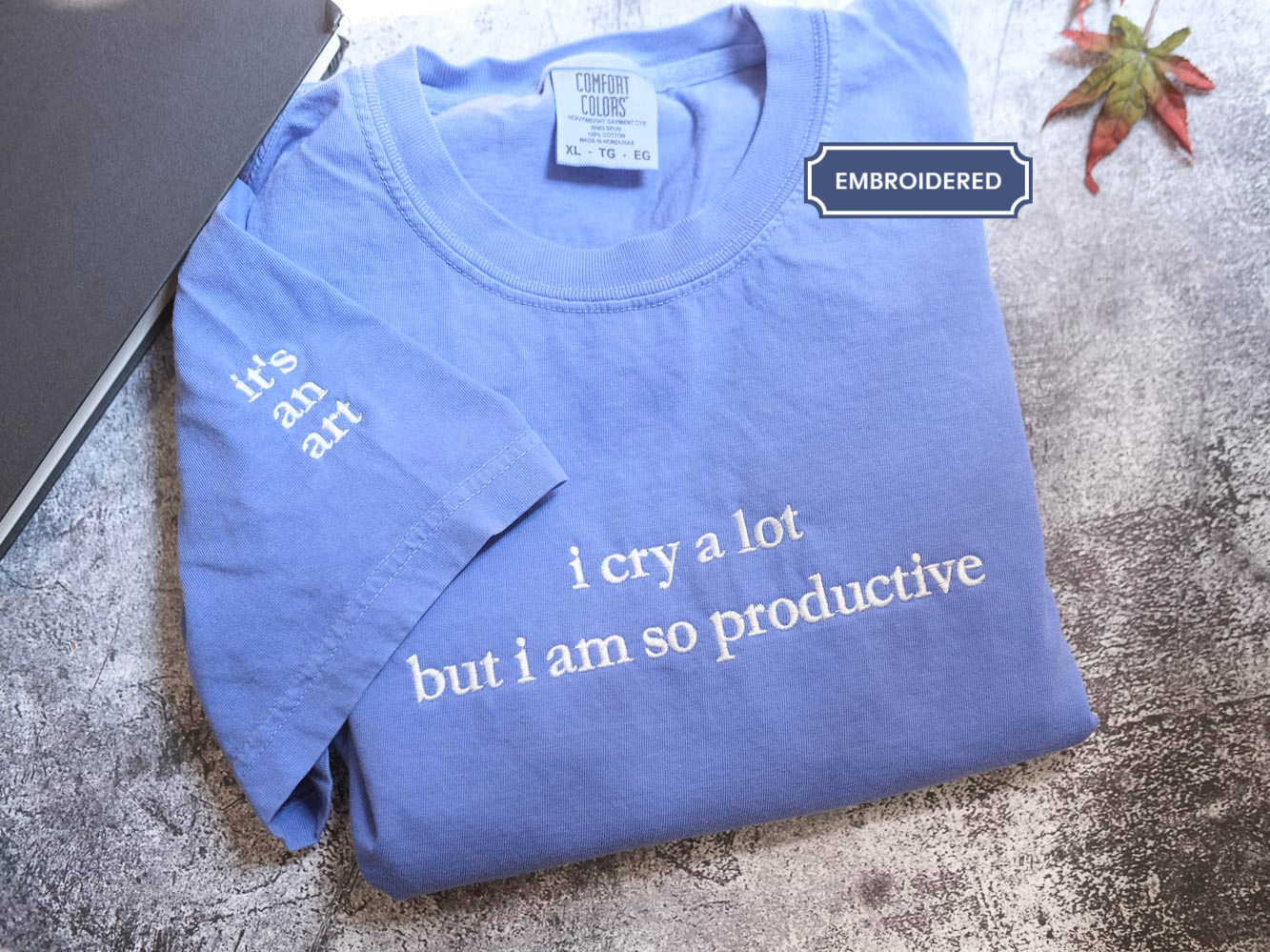 A blue t-shirt with white text is folded and placed on a gray surface, accompanied by a leaf and a laptop.