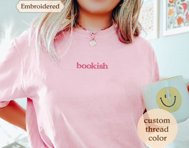 A person wearing a pink t-shirt with the word "bookish" embroidered on it, holding a mug with a smiley face design.