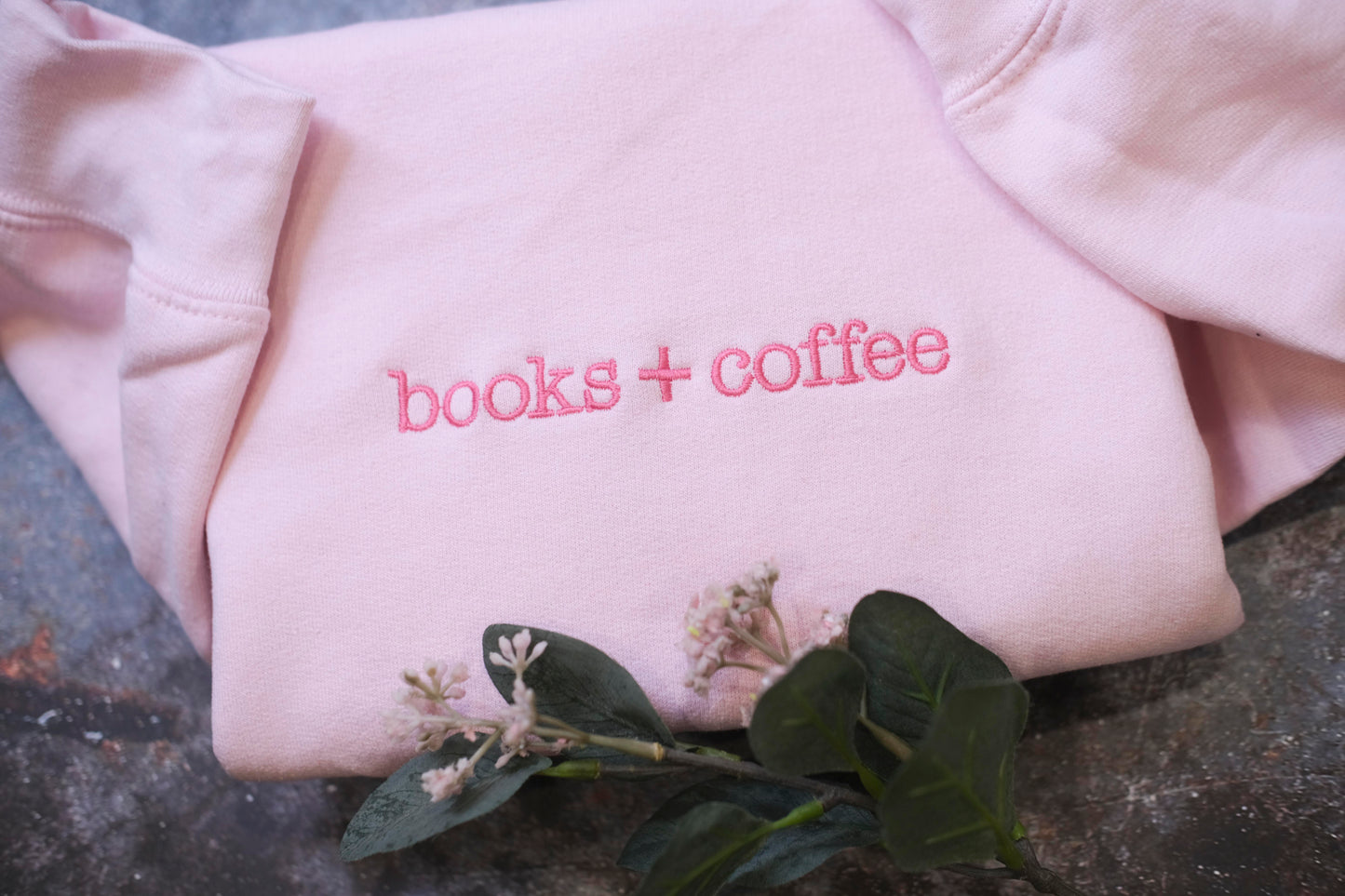 A pink sweatshirt with the text "books + coffee" embroidered on it, along with some flowers and leaves on the surface.