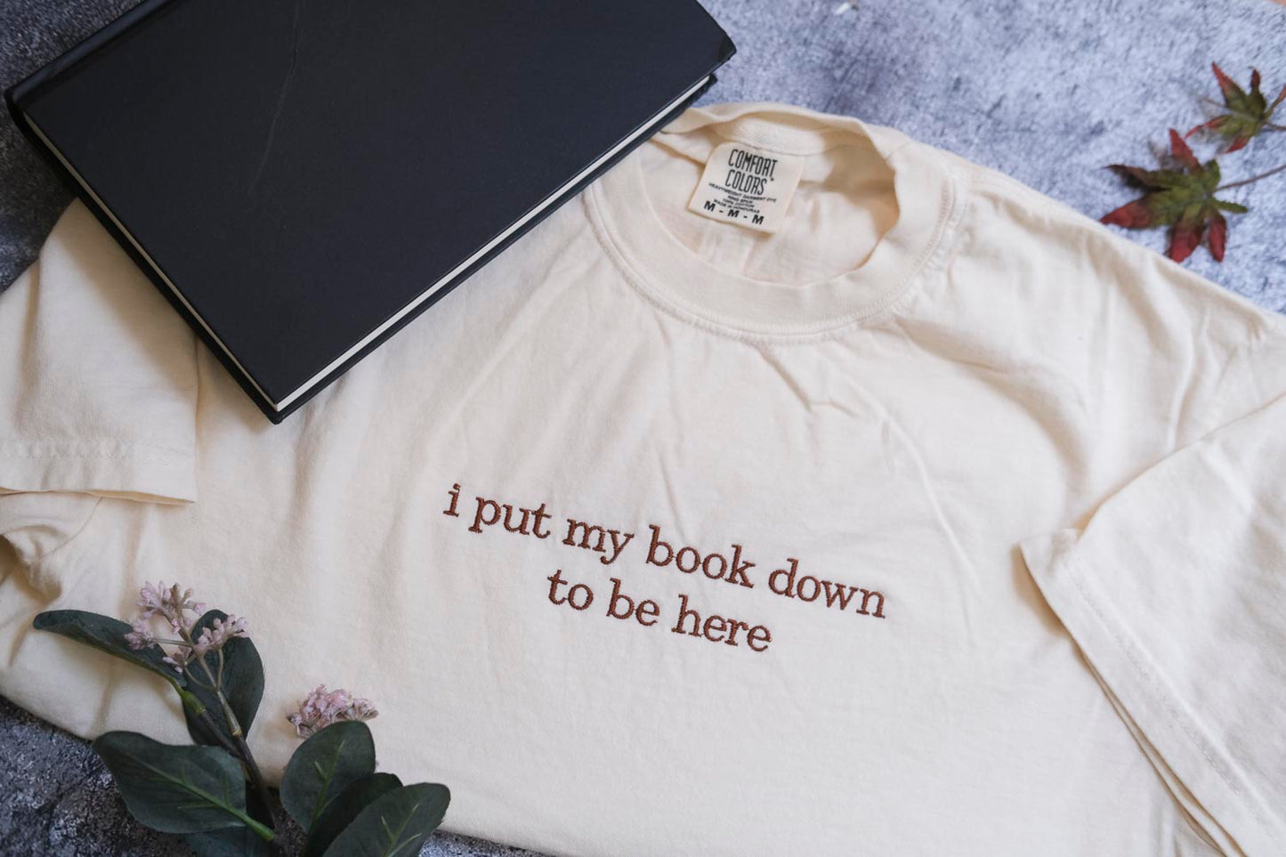 a white t-shirt with the text "I put my book down to be here" printed on it, along with a black notebook and some leaves.
