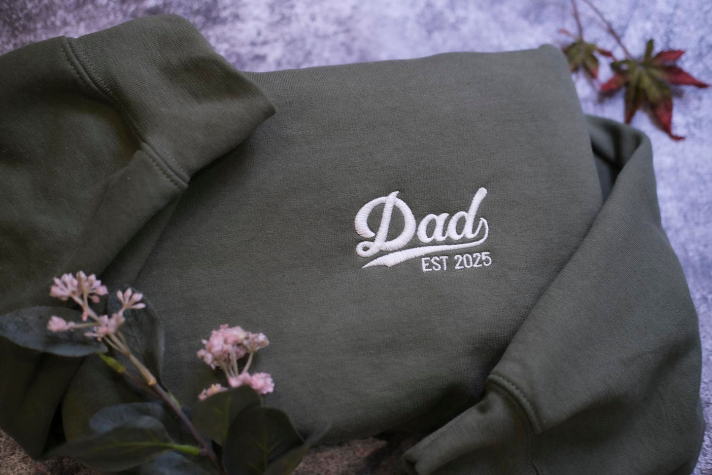A dark green sweatshirt with the text "DAD EST 2025" embroidered on it, along with some pink flowers and a leaf in the background.